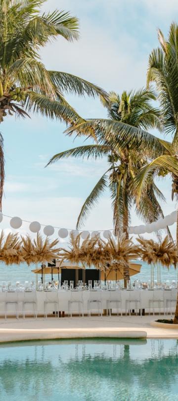 Une réception de mariage au style tropical organisée sur une plage, avec des palmiers, une longue table, des chaises et une piscine au premier plan.