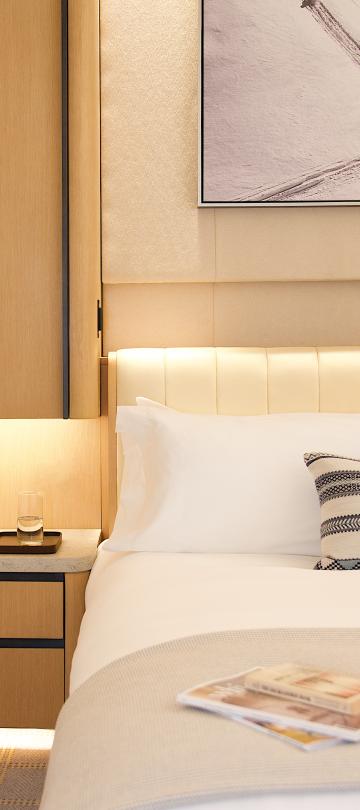 Hotel bed with white linens and a striped throw pillow next to a nightstand with books and water.