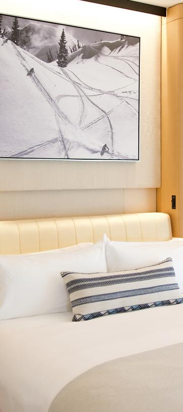 Angle view of a hotel bed with white linens, wooden side tables, and mountain artwork above.