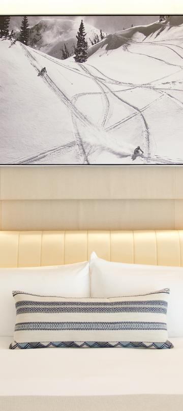 Cama de hotel moderna com roupa de cama branca, uma almofada decorativa estampada e, por cima, uma obra de arte emoldurada representando montanhas no inverno.