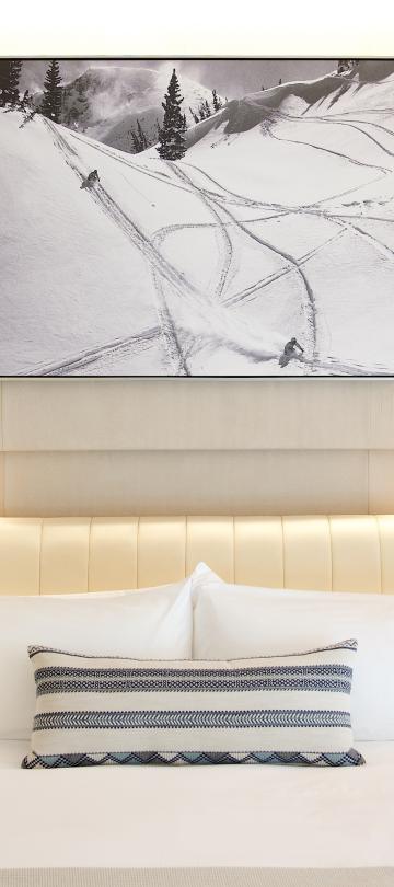 Hotel guest room bed framed by warm wooden built-in shelving with hanging clothing, books, and flowers.
