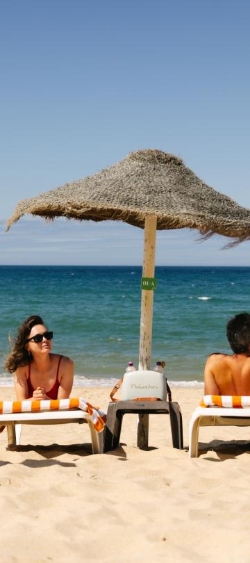 A family enjoys a day at the beach, with a girl playing with an orange ball and a boy standing near lounge chairs.