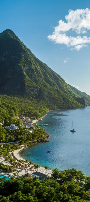 Des montagnes d'un vert luxuriant côtoient un littoral bordé par l'océan bleu, où naviguent des voiliers près de la plage et de la piscine d'un complexe hôtelier.
