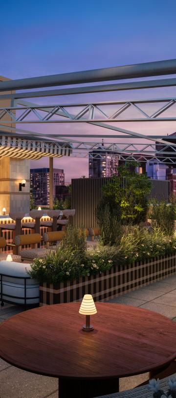 A rooftop restaurant patio at dusk has a bar, cushioned seating, round tables, planters, and a metal canopy overlooking city buildings.