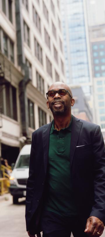 Um homem com óculos de sol, um blazer escuro e uma camisa verde caminha por uma rua da cidade, com edifícios altos e veículos estacionados atrás dele.