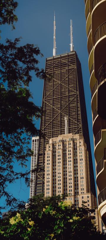 O John Hancock Center, um edifício escuro com fachadas em treliça, ergue-se entre árvores frondosas e um bloco de apartamentos curvo, com as suas antenas gémeas a destacarem-se contra o céu azul.