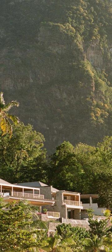 Un complexe touristique moderne, composé de bâtiments bas aux teintes claires, se niche au milieu d'une végétation tropicale, au pied d'une montagne escarpée et boisée.