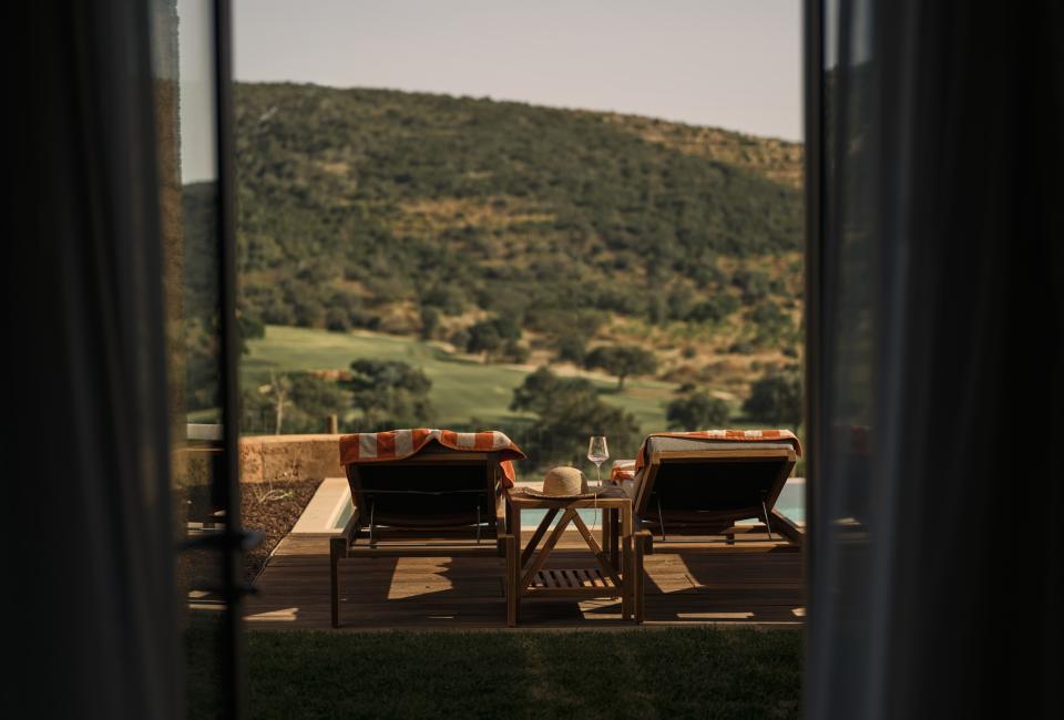 A view of two poolside recliners through a patio window, with the Algarve hills in the background