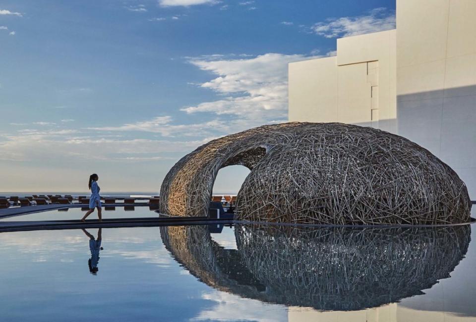 A woman walking on a boardwalk above water towards a dome made of branches.