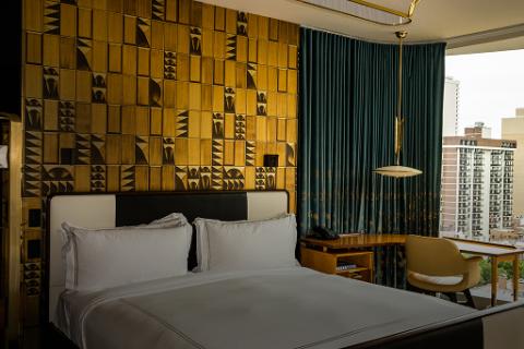 A room with a bed in white linen, gold decor on the wall, and dark blue curtains near a desk at a window.