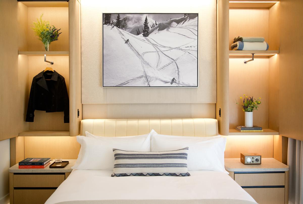 Hotel guest room bed framed by warm wooden built-in shelving with hanging clothing, books, and flowers.