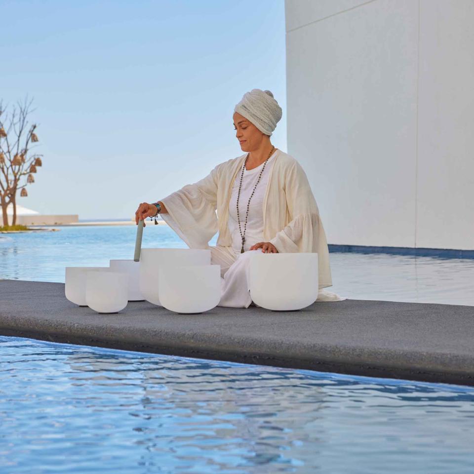 woman wearing white sits on platform in pool using sound bowls
