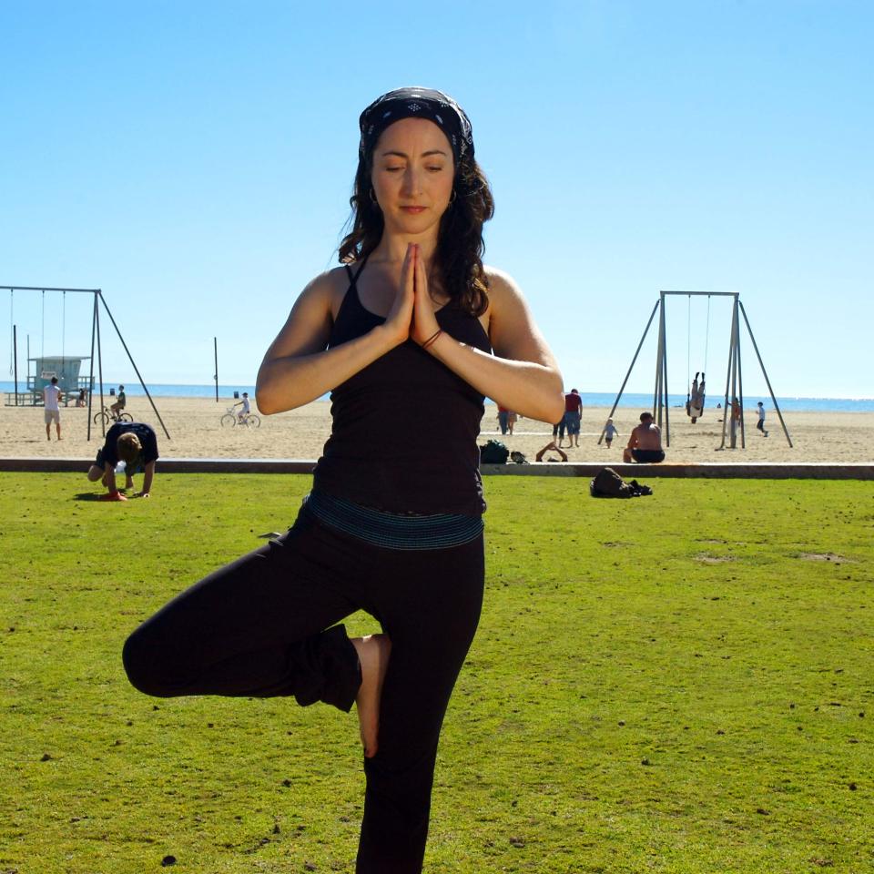 A woman holds a yoga pose, with her palms pressed together and one leg raised, near a playground