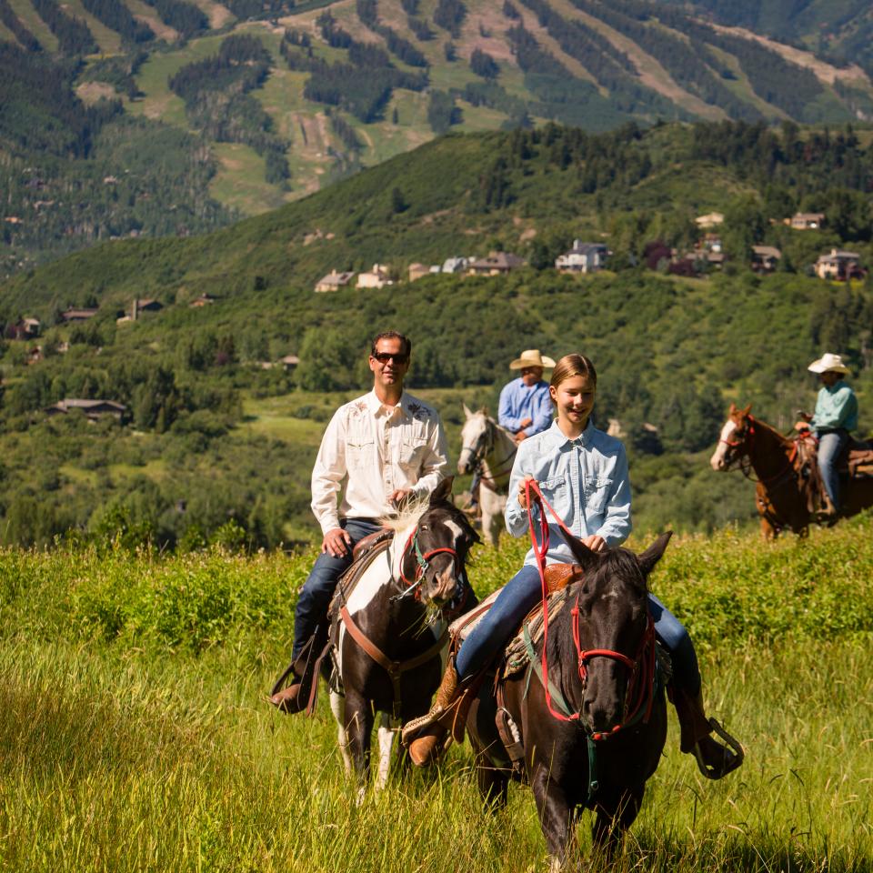 A young girl and a man ride horses through a lush green valley at the base of a mountain