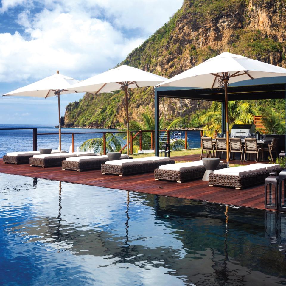 A row of pool recliners under patio umbrellas next to a pool, in view of the Pitons at Sugar Beach