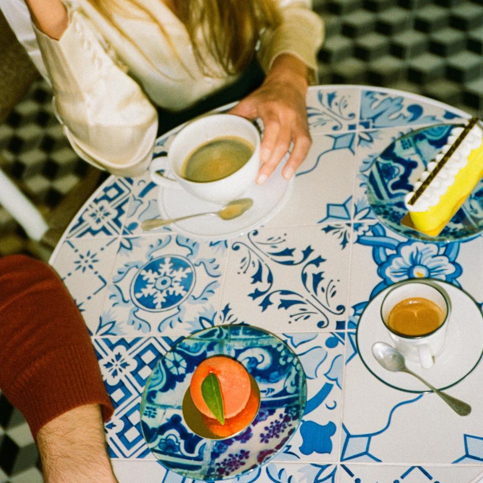 Dos personas sentadas ante una mesa con un estampado azul, tomando café y postres