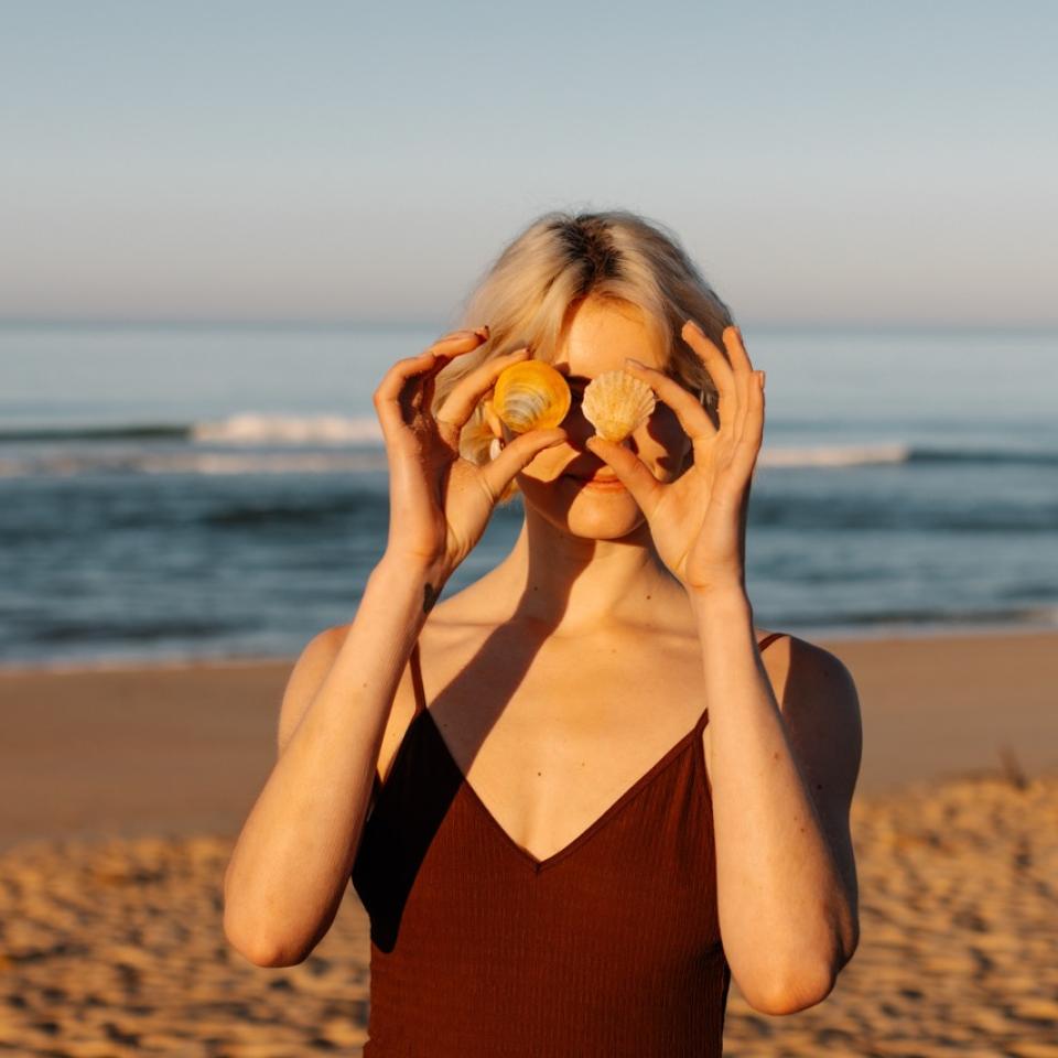 A woman holds sea shells in front of her eyes like sunglasses, as she stands with her back to the sea.