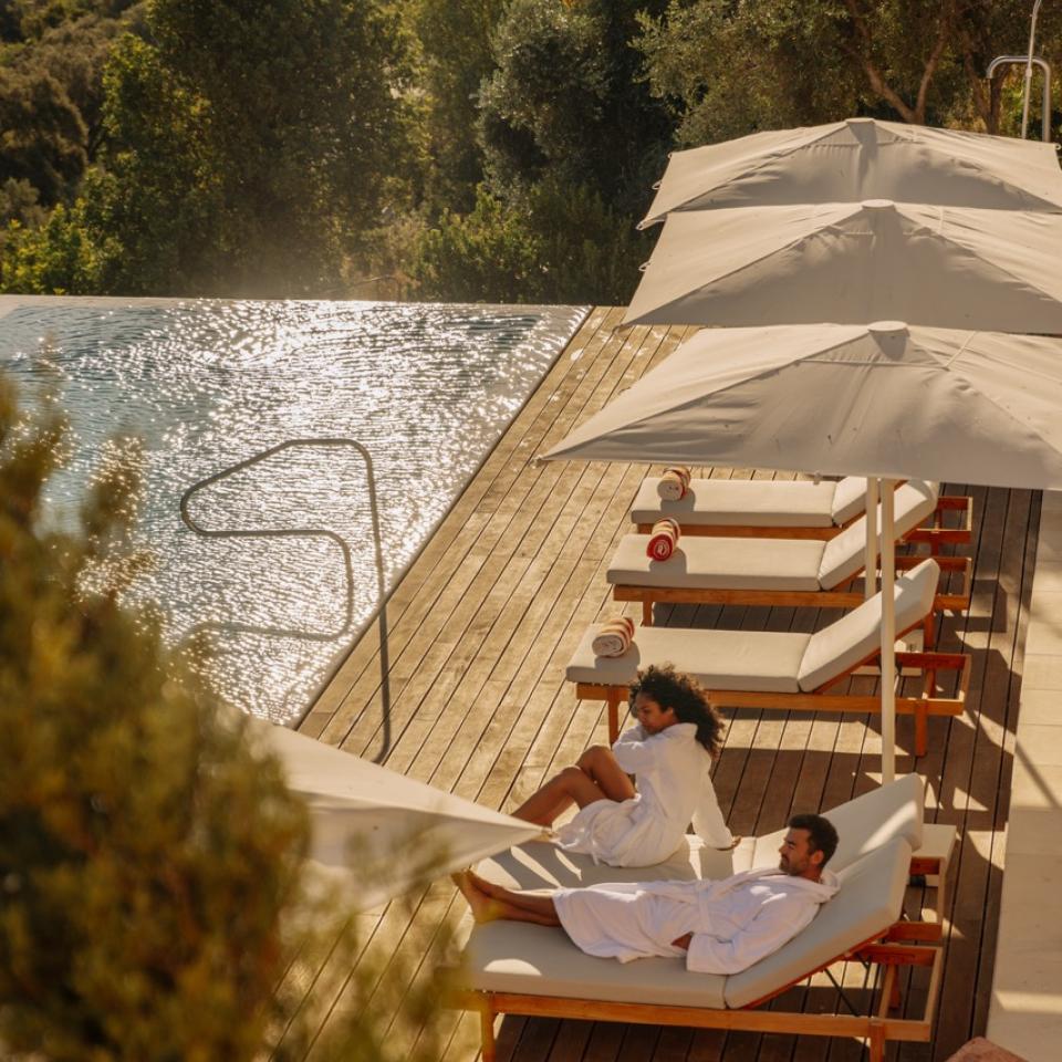 A couple relaxing on loungers under an umbrella beside the pool.