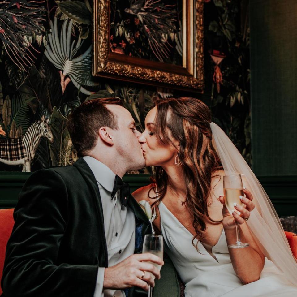 A bride and groom sitting in a red restaurant booth, kissing, holding flutes of champagne