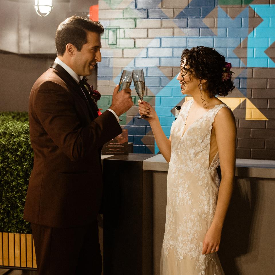A smiling groom in a brown suit and a bride in a white lace gown toast with champagne flutes in front of a colorful geometric brick wall.