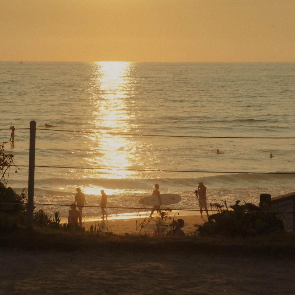 Au coucher du soleil, on aperçoit au bord de la mer les silhouettes de baigneurs et d'un surfeur portant sa planche, tandis que le soleil se reflète avec éclat sur l'eau de la mer.