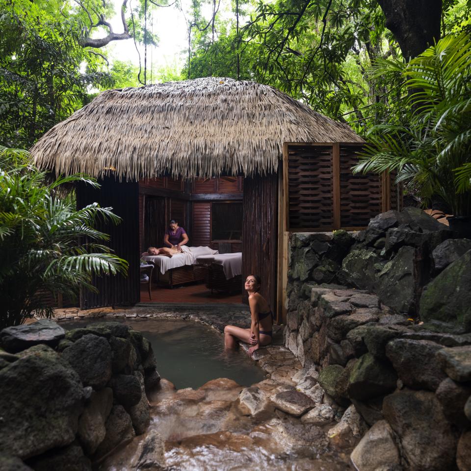 Une femme est assise au bord d'une source thermale naturelle, à côté d'un spa au toit de chaume où l'on procède à un massage.