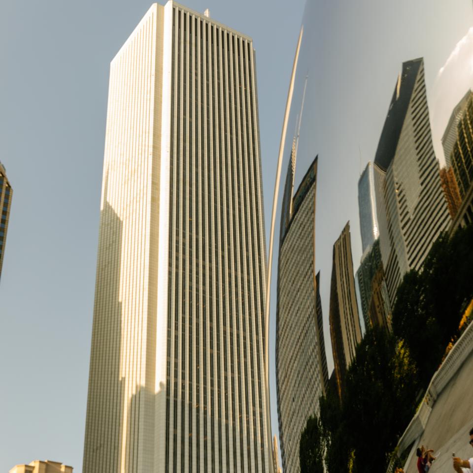 Un rascacielos alto y blanco reflejado en una escultura curvada y reflectante, con personas en primer plano.