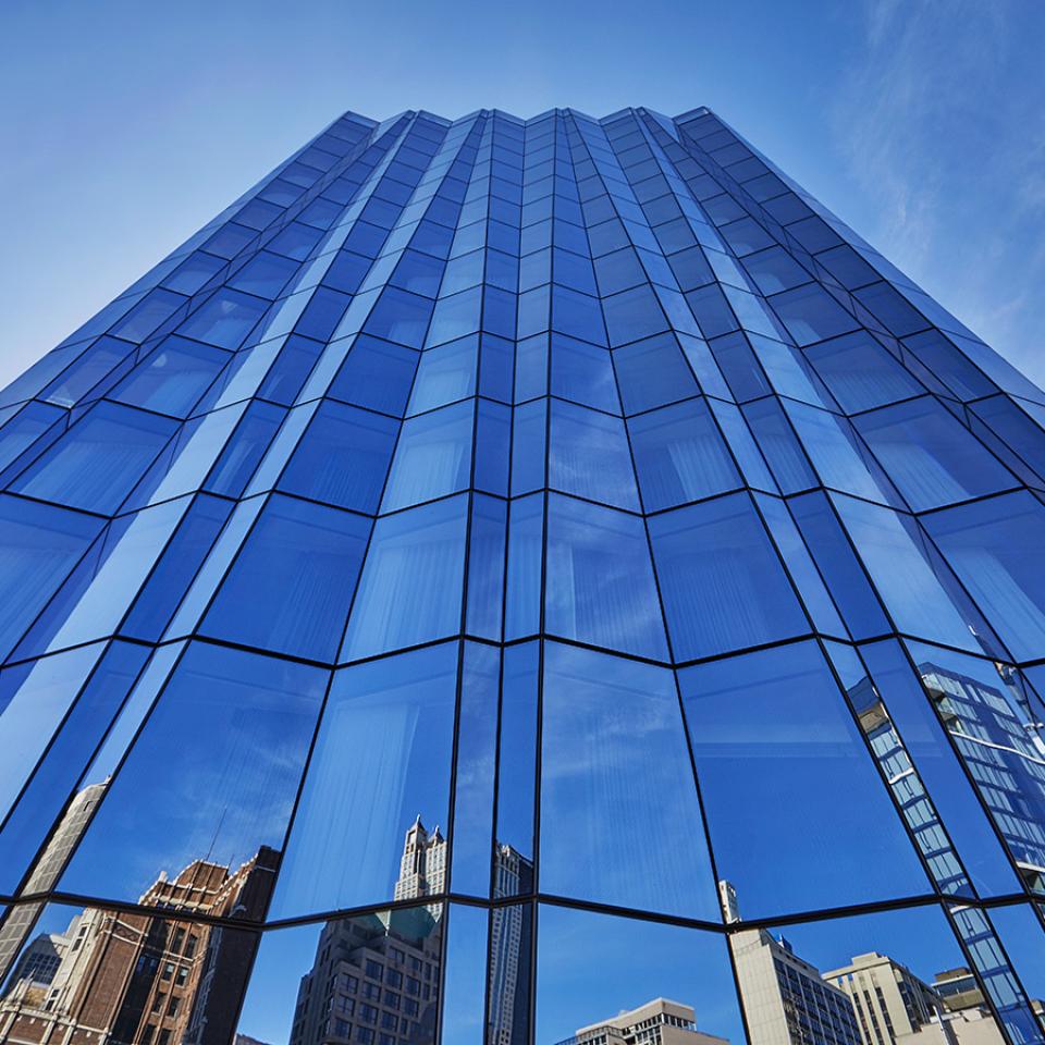 Vista desde un ángulo bajo de un rascacielos moderno con fachada de cristal azul en la que se reflejan los edificios y el cielo.
