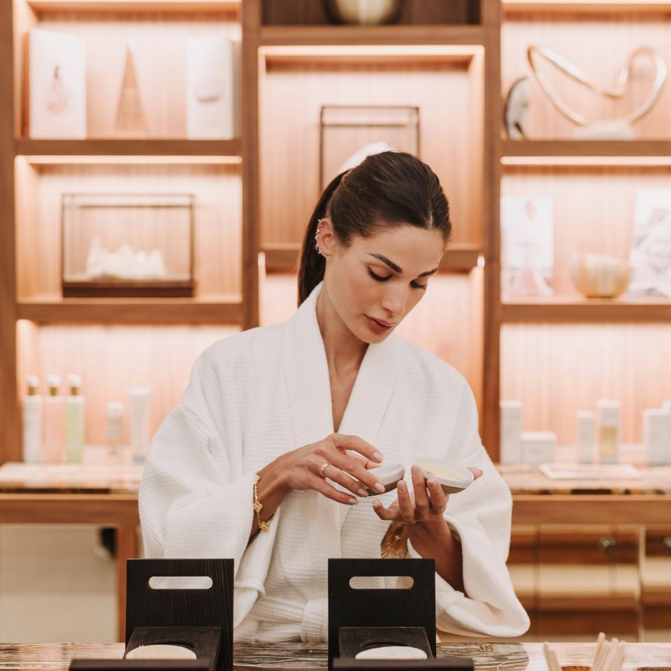 Une femme vêtue d'une robe blanche examine un petit flacon de cosmétique devant des rayons remplis de produits de soin pour la peau.