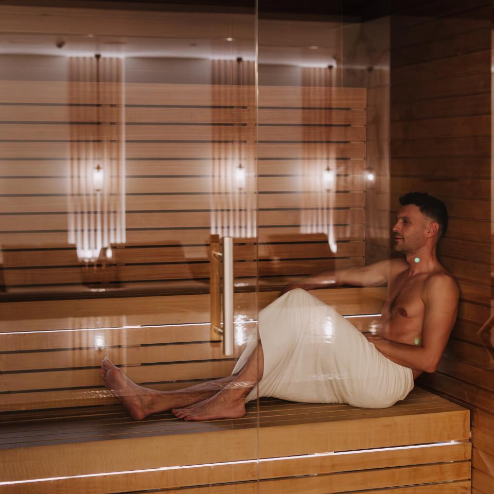 Un homme, vêtu d'une serviette blanche, se détend sur un banc en bois dans un sauna moderne.