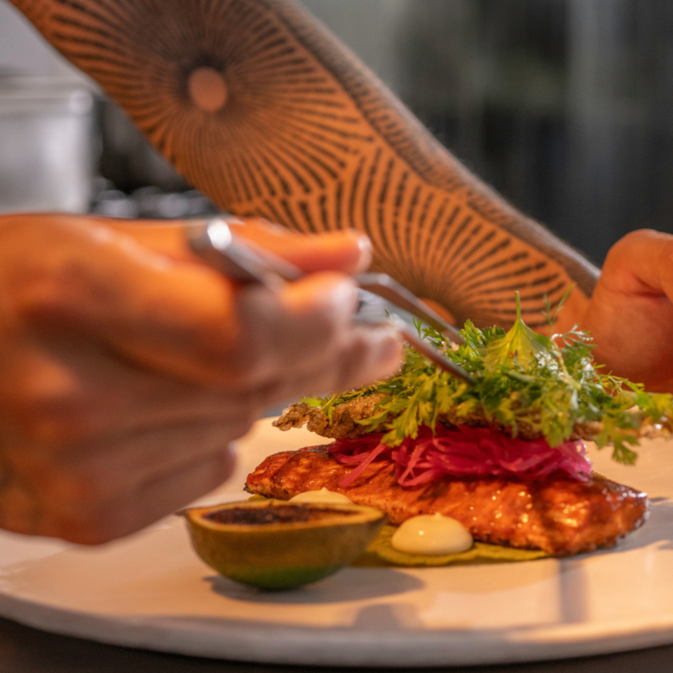 Le chef décore un plat à l'aide d'une pince à cuisine, en dressant un filet de saumon accompagné d'oignons marinés et de micro-pousses.