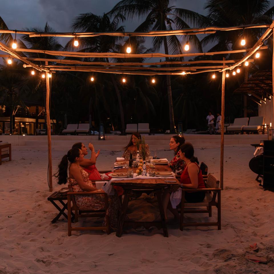 À la tombée de la nuit, cinq personnes dînent à une table sur une plage de sable, sous un dais formé de guirlandes lumineuses.