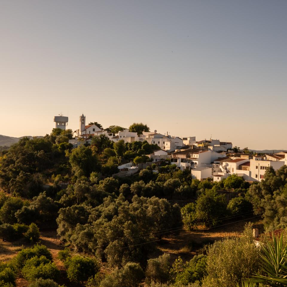 Elevated wide view of a small Portuguese village with white houses, surrounded by green trees and hills under a warm, hazy sky.