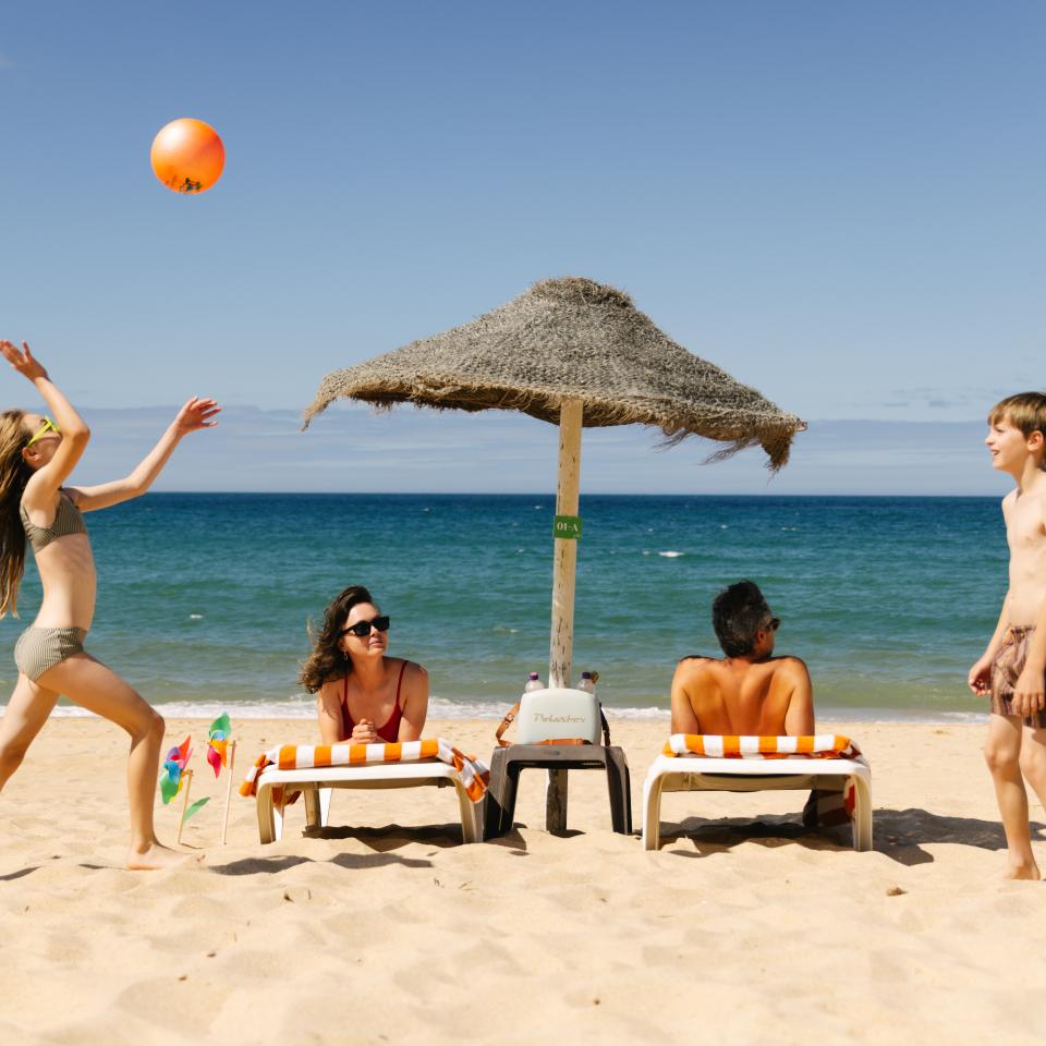 A family enjoys a day at the beach, with a girl playing with an orange ball and a boy standing near lounge chairs.