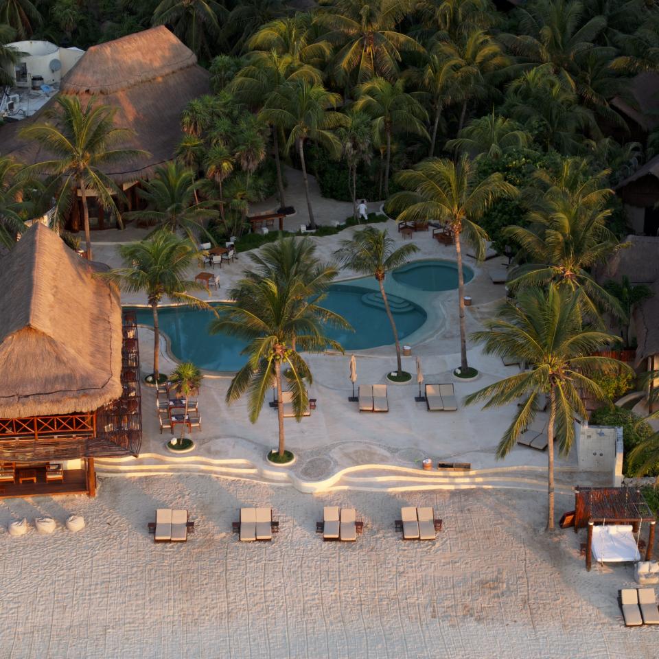 Aerial view of a tropical beach resort with thatched-roof huts, palm trees, a turquoise swimming pool, and lounge chairs on white sand.