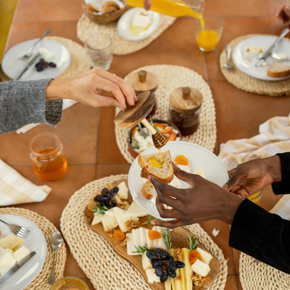 People share a brunch table with toast, honey, orange juice, yogurt, fruit, cheese, and charcuterie served on woven placemats.