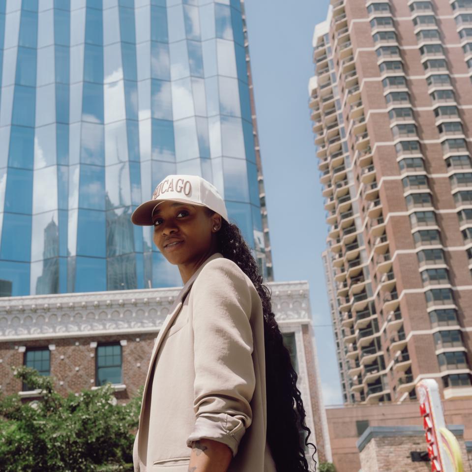 Uma mulher com um casaco bege e um boné com a inscrição «CHICAGO» encontra-se entre os edifícios do centro da cidade, com uma torre de vidro e um arranha-céus atrás dela.
