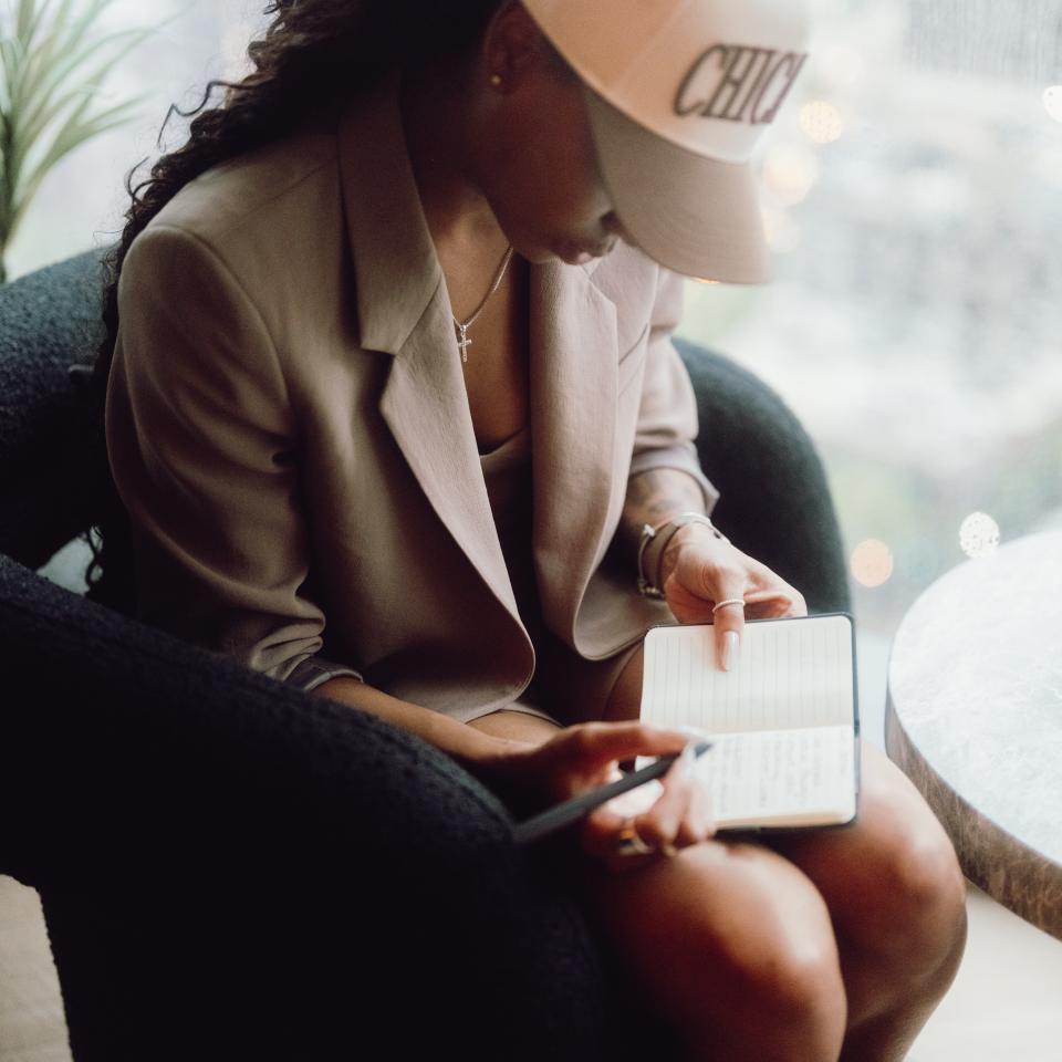Uma mulher, vestida com um blazer bege e um boné branco, está sentada junto a uma janela, com o olhar voltado para baixo enquanto escreve num caderno aberto.