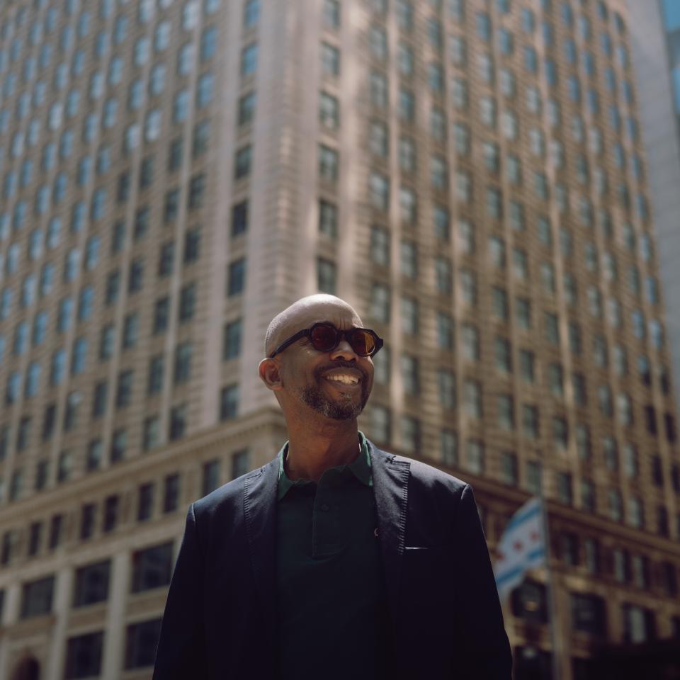 Um homem careca, sorridente, com óculos de sol e um casaco escuro, está em frente a um edifício alto de pedra, com janelas ao longo de toda a fachada.
