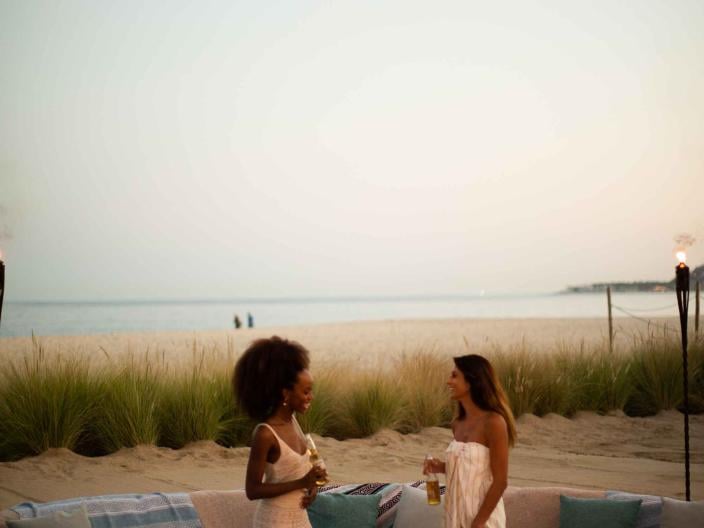 two women stand near a firepit surrounded by coaches on the beach