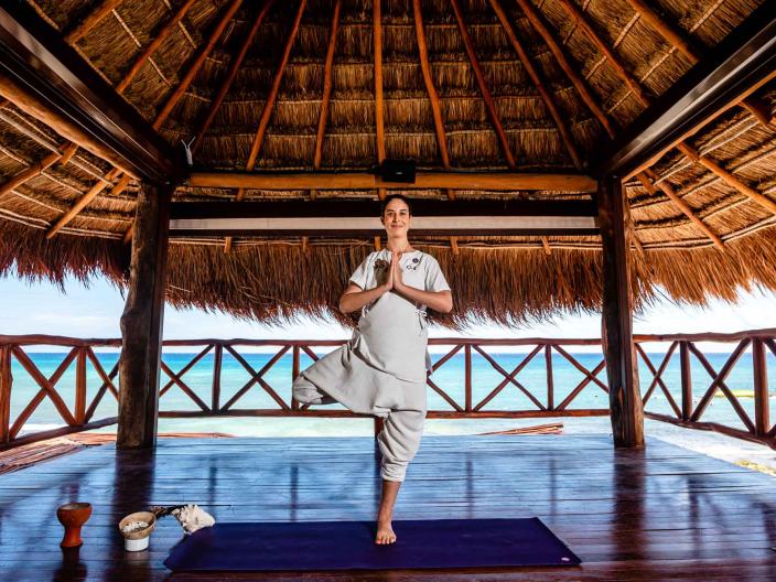 woman stands in a yoga pose thatched roof hut on beach