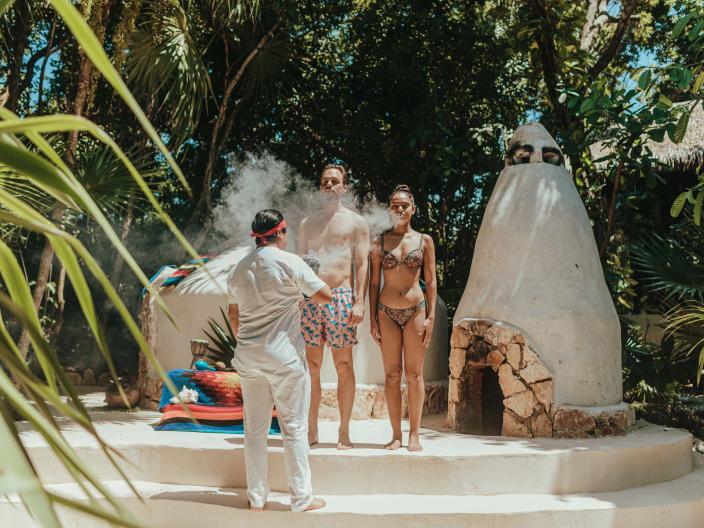 couple in bathing suits stand on a altar while a person wafts smoke towards them
