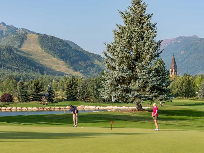 two people play golf with mountains and trees in background