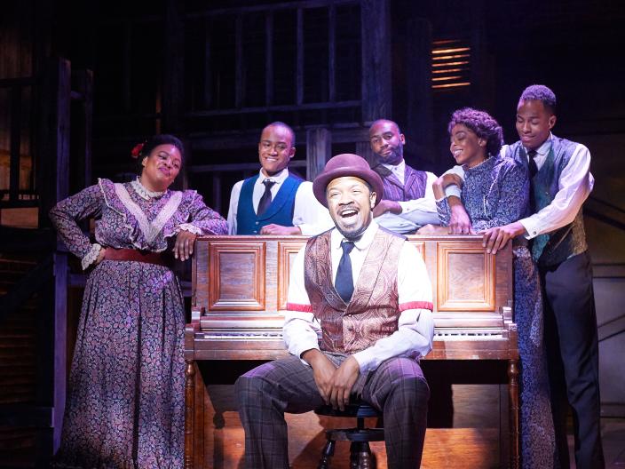 group of actors around a piano dressed in suits and ties