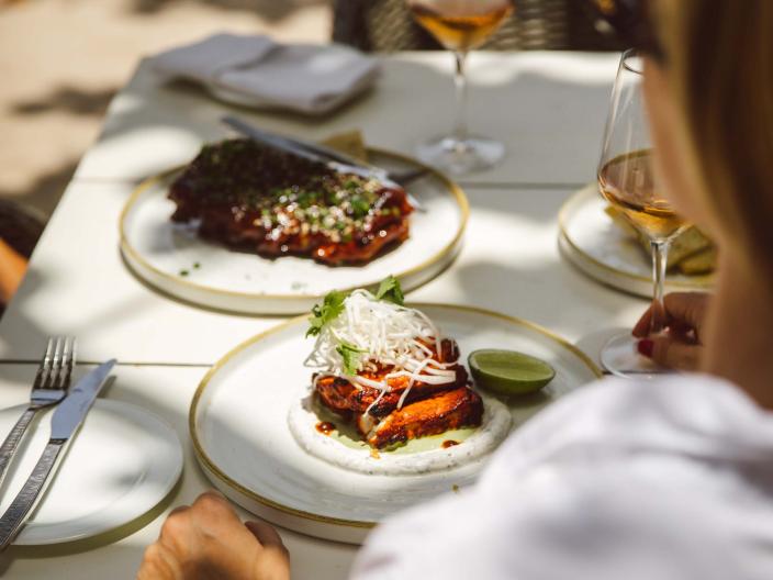 woman wearing white shirt sits at table on beach with plates of food