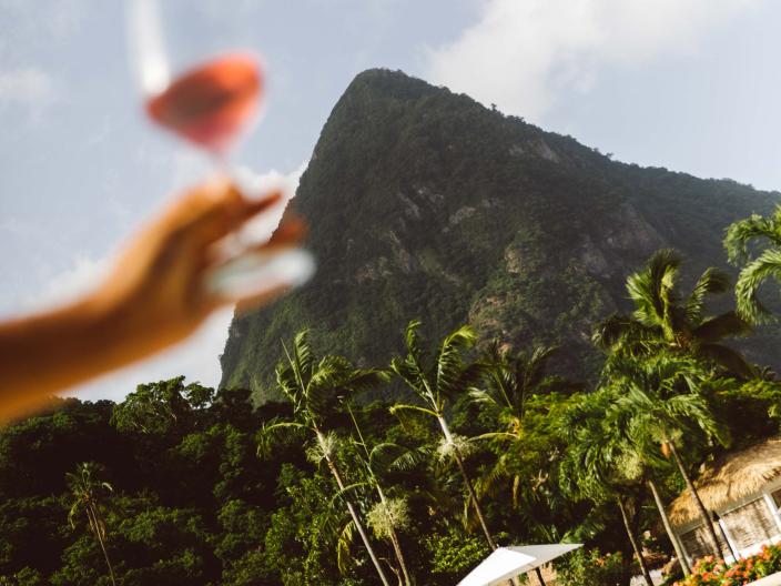 person holding wine glass in front of pool, cabanas, and mountain