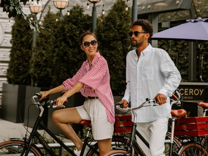 A couple rides bicycles with red baskets on the back, in downtown Chicago near the Viceroy Chicago entrance