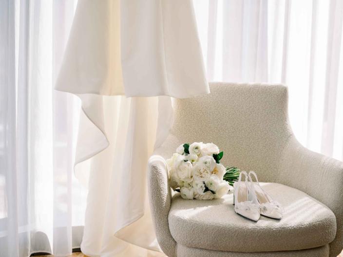 A veil hung up near a white armchair with a bridal bouquet and shoes resting upon it, near a white curtain in a light-streaked hotel room