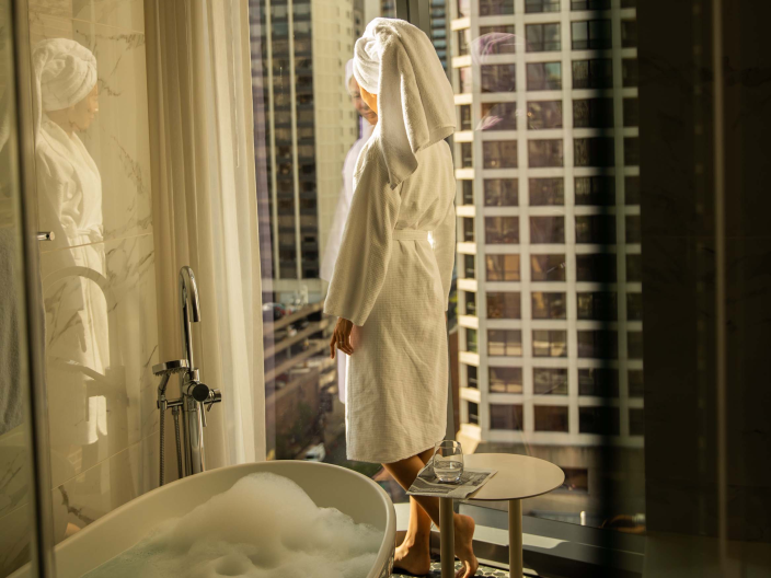 A woman standing in the bathroom in a housecoat staring out the window at Viceroy Chicago