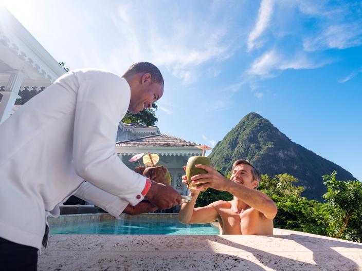 A Viceroy staff member brings a drink served in a coconut to a guest in a hot tub, in a tropical locale at the foot of a mountain